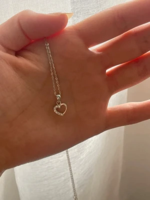 Äkta silverhalsband  - Säljer ett tunt silverfärgat halsband med ett litet hjärthänge dekorerat med små gnistrande pärlor på ena sidan. Kedjan är enkel och stilren, perfekt för dig som gillar minimalistiska smycken med en romantisk touch.
