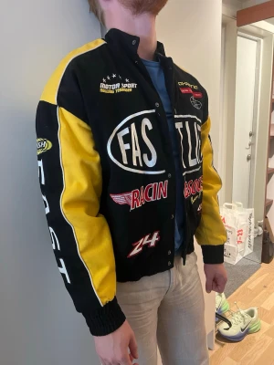 Svart och gul racingjacka med tryck - Säljer en svart och gul racinginspirerad jacka med stora tryck och broderade detaljer på bröst, ärmar och rygg. Jackan har tryckknappar framtill, muddar vid ärmslut och nederkant samt coola racingloggor som 'FAST', 'FLASH' och 'RACIN'. Perfekt för dig som gillar streetwear.
