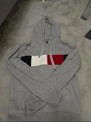 Grå hoodie från Tommy Hilfiger - Snygg grå hoodie från Tommy Hilfiger med klassisk logga i blått, vitt och rött på bröstet. Tröjan har huva med dragsko och ribbade muddar vid ärmar och nederkant. Perfekt för en chill och stilren look.