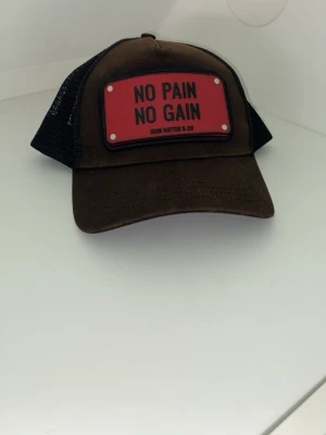 Brun truckerkeps John Hatter & Co - Snygg svart men lite solblekt truckerkeps från John Hatter & Co med svart mesh baktill och böjd skärm. Framtill sitter en röd patch med texten 'NO PAIN NO GAIN'. Kepsen har justerbar snapback och är tillverkad i bomull och polyester.