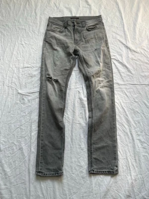 Grå jeans från Nudie Jeans Co - Säljer ett par grå jeans från Nudie Jeans Co i modellen Lean Dean. Jeansen har en slim fit, klassisk femficksdesign och ett litet slitmärke på vänster lår. Tillverkade i mjuk denim med snygg tvättad look.
