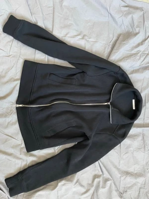 Svart zip up hoodie från ZARA - Snygg svart zip up hoodie från ZARA med ribbade muddar, hög krage och dragkedja framtill. Hoodien har två fickor på sidorna och är tillverkad i ett mjukt material som känns bekvämt. Perfekt för en stilren och enkel look.