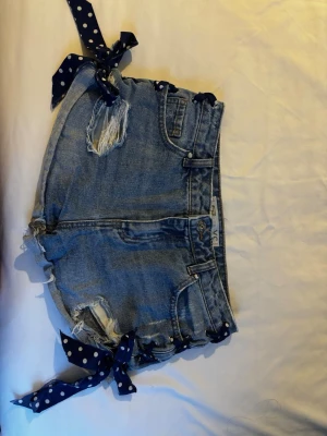Blå jeansshorts med prickiga knytband - Säljer ett par blå denimshorts med slitna detaljer och coola knytband i midjan och benen. Knytbanden är mörkblå med vita prickar som ger en unik touch. Shortsen har klassisk femficksdesign och råa kanter nertill. Perfekta för sommaren!