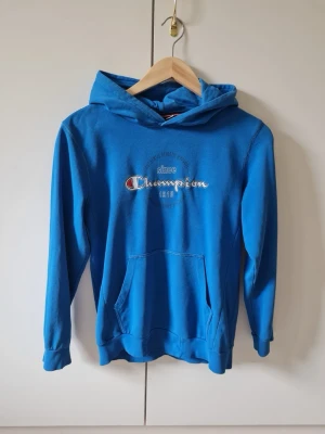 Blå hoodie från Champion strl 150/155 cm 11/12 yrs barn - Snygg blå hoodie från Champion med klassisk logga broderad på bröstet. Hoodien har huva, känguruficka framtill och ribbade muddar vid ärmslut och nederkant. Material 85% bomull, 15% polyester. Storlek 150/155 cm, 11/12 yrs.