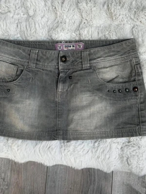 Grå kort jeanskjol med nitar - Säljer en grå kort jeanskjol med slitningar och coola nitar på fickorna. Kjolen har bälteshällor, knappstängning och bakfickor med dekorativa sömmar. 