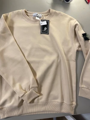 Beige sweatshirt från Stone Island - Snygg beige sweatshirt från Stone Island med klassisk rund halsringning och ribbade muddar. Tröjan har den ikoniska Stone Island-loggan på vänster ärm och är tillverkad i mjukt material som känns skönt mot huden. Lapparna är ej kvar. Storlek M men passar nog bättre på S