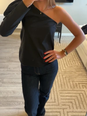 Svart oneshoulder topp - Snygg svart topp med oneshoulder-design och lång ärm på ena sidan. Tidlös och stilren modell som passar perfekt till jeans eller kjol. Materialet känns mjukt och följsamt mot huden.