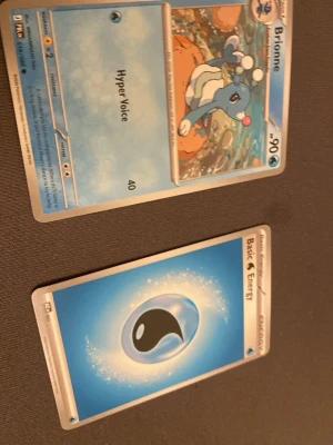 Två Pokémonkort: Brionne & Vattenenergi - Säljer två Pokémonkort, ett med Brionne och attacken Hyper Voice samt ett Basic Water Energy-kort. Båda korten har blå och vita färger och klassisk Pokémon-design. Perfekt till samlingen eller för att boosta din lek.