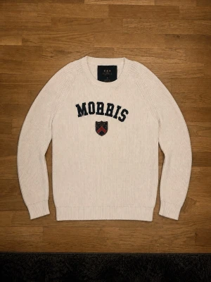 Vit stickad tröja från Morris - Säljer en stilren vit stickad tröja från Morris med text och emblem på framsidan. Tröjan har långa ärmar och en rund halsringning. Perfekt för en klassisk och avslappnad look.