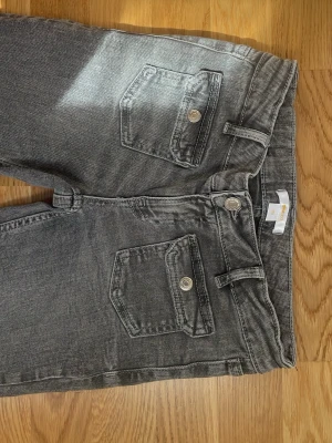 Grå jeans från Gina Tricot Young  - Snygga grå jeans från Gina Tricot med två framfickor med lock och knappdetaljer. Klassisk femficksmodell med bälteshällor och dragkedjegylf. Jeansen har en tvättad look och är tillverkade i mjukt denimtyg. Storlek 152. Två små fläckar.