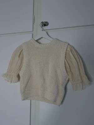 Vit stickad väst med puffärmar - Säljer en krämvit stickad väst med bred ribbad kant och rund hals. Under västen syns en blus med puffiga ärmar och volangdetaljer vid ärmsluten. Perfekt för lager på lager och ger en romantisk, vintage-inspirerad vibe.