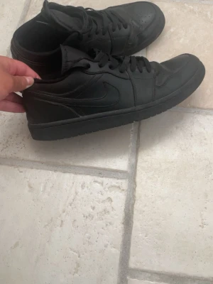 Helsvarta låga skor - Snygga helsvarta sneakers med lågt skaft och klassisk siluett. Skorna har en stilren design med matchande svarta skosnören och sula. Tillverkade i skinnliknande material och har perforeringar på tån för extra ventilation.