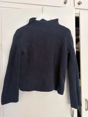 Mörkblå ribbstickad tröja - Mysig mörkblå ribbstickad tröja med hög krage och långa ärmar. Tröjan har en rak passform och är tillverkad i ett tjockare garn som ger en skön och avslappnad look. Perfekt för kyliga dagar och enkel att matcha med jeans eller kjol.