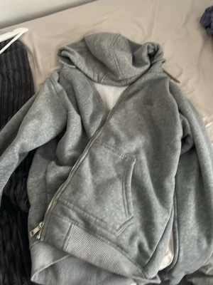 Grå hoodie med mönstrat foder - Säljer en grå hoodie med dragkedja och huva. Insidan har ett mjukt vitt foder och klassiskt rutigt mönster i beige, svart och rött. Hoodien har fickor framtill och ribbade muddar vid ärmslut och nederkant. Perfekt för en avslappnad och stilren look.