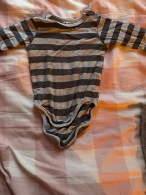 Randig body från H&M i ekologisk bomull - Mjuk och bekväm långärmad body från H&M i ekologisk bomull. Grå och mörkgrå breda ränder, rund hals och tryckknappar i grenen för enkel påklädning. Perfekt basplagg för små barn.