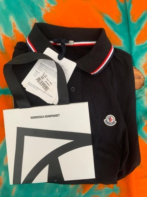 Svart pikétröja från Moncler - Snygg svart pikétröja från Moncler med klassisk krage och rödvit rand på kragen. Tröjan har knappslå framtill och Moncler-logga broderad på bröstet. Perfekt för en stilren och sportig look.