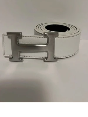Vitt bälte med stor H-spänne - Snyggt vitt bälte med ett stort, blankt H-format metallspänne. Bältet har en stilren och modern look med tydliga sömmar längs kanterna. Perfekt accessoar för att lyfta vilken outfit som helst.