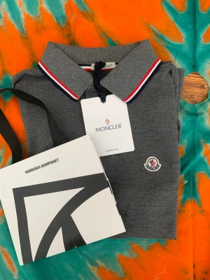 Grå pikéskjorta från Moncler XS - Säljer en grå pikéskjorta från Moncler i storlek XS. Skjortan har klassisk krage med röd, vit och marinblå rand, samt Moncler-logga på bröstet. Tillverkad i mjuk bomull och perfekt för en clean och stilren look.