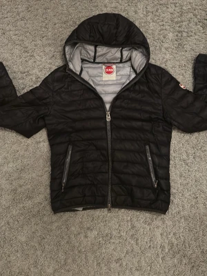 Colmar jacka - Snygg svart jacka från Colmar med quiltad design och huva. Jackan har två dragkedjeförsedda fickor framtill och grått innerfoder. Jackan är i storlek 50 som sitter som S.  Perfekt för vintern och hösten och enkel att matcha till olika outfits.
