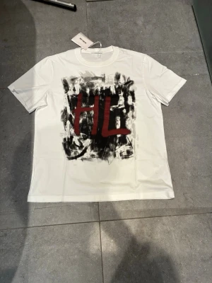 Vit Helmut Lang t-shirt med tryck - Säljer en vit t-shirt från Helmut Lang med coolt grafiskt tryck i svart och rött både fram och bak. Framsidan har stora bokstäver 'HL' och baksidan har Helmut Lang-loggan. Klassisk passform och rund hals. Perfekt för dig som gillar streetwear vibes.