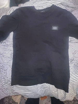 Svart sweatshirt från Dolce & Gabbana - Säljer en svart sweatshirt från Dolce & Gabbana med en snygg metallbricka på bröstet där märket står. Tröjan har rund halsringning och ribbade muddar vid ärmslut och nederkant. Perfekt för dig som gillar stilrena och exklusiva plagg.
