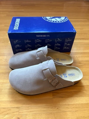 Birkenstock Boston ljusgrå tofflor - Säljer ett par snygga ljusgrå Birkenstock Boston i storlek 42. Som nya och i mycket fint skick utan några brister.  Klassisk och bekväm modell i mjuk mocka med justerbart metallspänne och ergonomisk fotbädd i kork. Perfekta både hemma och ute!  Originalkartong följer med.  Vid bud eller funderingar är det bara att skriva! 😊