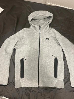 Grå Nike tech - Snygg ljusgrå hoodie från Nike med svart logga på bröstet. Hoodien har hel dragkedja framtill, två svarta dragkedjeförsedda fickor och en stor huva. Tillverkad i mjukt material som känns skönt mot huden. Perfekt för en sportig och avslappnad stil.