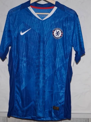 Chelsea FC blå matchtröja Nike - Chelsea FCs officiella blå matchtröja från Nike med klubbmärke på bröstet och vit Nike-logga. Tröjan har korta ärmar, V-ringning med vit och röd detalj samt ett mönstrat tyg i polyester som andas. Perfekt för fotbollsträning eller att bära på läktaren. Är du efter en annan storlek är det bara att skriva!