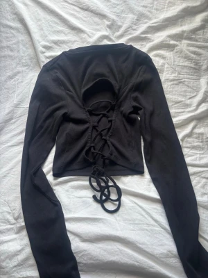 Svart croppad topp med öppen rygg och snöring  - Trendig svart croppad topp med långa ärmar och snygg öppen rygg med snöring. Tillverkad i ribbad bomull som ger en skön och stretchig känsla. Perfekt för dig som vill ha en edgy och modern look. Från New yorker i storlek M men skulle passa bra för dig som är S,XS eftersom den är ganska liten i storlek.💕💕