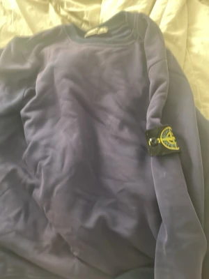 Blå sweatshirt från Stone Island - Säljer en blå sweatshirt från Stone Island med den klassiska logotypen på vänster ärm. Tröjan har rund halsringning och är tillverkad i mjukt material, perfekt för en avslappnad och stilren look.