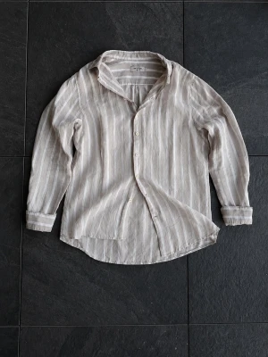 Randig linneskjorta från Dressmann - Säljer en snygg randig skjorta från Dressmann i beige och vitt. Skjortan är tillverkad i linne, har långa ärmar och klassisk krage. Perfekt för dig som gillar en avslappnad men stilren look. Stängs med knappar framtill.