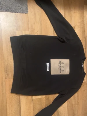 Svart sweatshirt från Burberry - Stilren svart sweatshirt från Burberry med beige patch och tydlig logga på bröstet. Tröjan har rund halsringning och långa ärmar. Perfekt för dig som gillar klassisk design med en modern twist.
