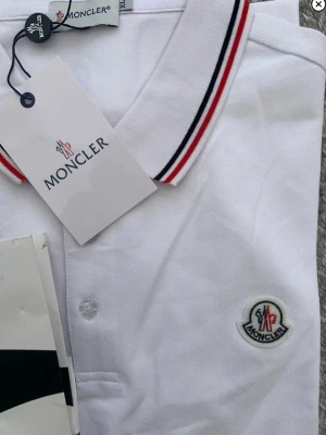 Vit pikétröja från Moncler XL - Säljer en fräsch vit pikétröja från Moncler i storlek XL. Tröjan har klassisk krage med röda och svarta ränder, knappar framtill och Moncler-logga broderad på bröstet. Tillverkad i bomull för en skön och luftig känsla.