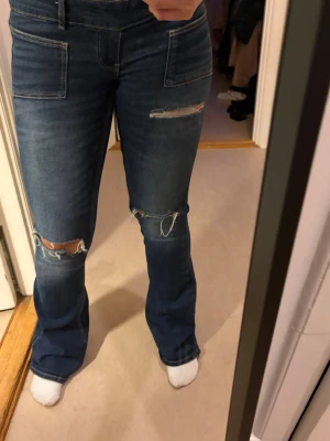 Blå bootcut jeans med slitningar - Säljer ett par blå bootcut jeans med råa slitningar på låret och knät. Jeansen har hög midja, stora framfickor och är i ett stretchigt denimtyg. Perfekta för en avslappnad och trendig look.