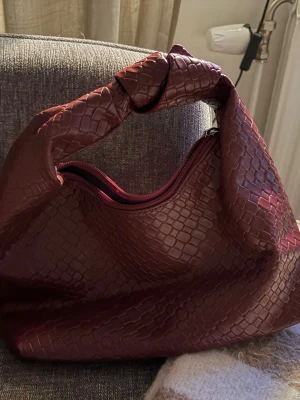 Vinröd handväska med knutdetalj - Säljer en vinröd handväska i fejkläder med ormmönstrad struktur. Väskan har en unik knutdetalj på handtaget och stängs med dragkedja. Perfekt för dig som vill ha en trendig accessoar med lite extra edge.