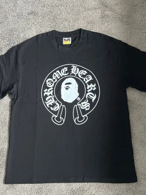 Svart Chrome Hearts x Bape t-shirt - Säljer en svart t-shirt från Bape med stor Chrome Hearts-inspirerad print på bröstet. Trycket är vitt och har en ikonisk Bape-apa i mitten omgiven av texten 'Chrome Hearts' i gotisk stil. Klassisk rund hals och korta ärmar.