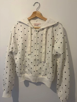 Vit hoodie med svarta prickar och text - Säljer en vit hoodie med svarta prickar och dragkedja framtill. På ryggen står det 'Obsessed' i röd text. Hoodien har huva med snören och fickor framtill. Perfekt för dig som gillar en lekfull och trendig stil.