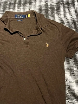 Brun pikétröja från Polo Ralph Lauren - Snygg brun pikétröja från Polo Ralph Lauren med klassisk krage och två knappar framtill. Tröjan har det ikoniska broderade logotypen på bröstet och är tillverkad i mjuk bomull. Perfekt för en avslappnad men stilren look.