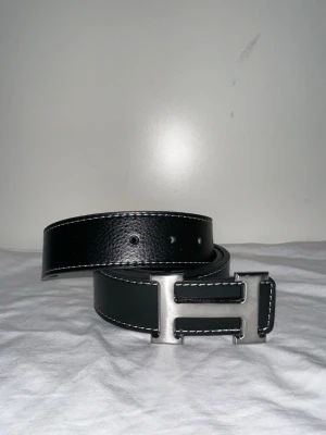 Svart bälte med silverfärgat spänne - Snyggt svart bälte i skinn med vit kontrastsöm och ett stort, silverfärgat H-format spänne. Perfekt accessoar för att lyfta din outfit med en stilren och modern touch. Bältet har klassisk design och passar till många olika looks.