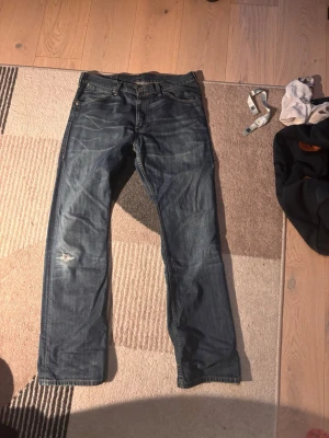 Blå raka jeans från Levi's - Säljer ett par klassiska blå jeans från Levi's med raka ben och fem fickor. Jeansen har slitningar på vänster lår och tydliga sömmar baktill. Tillverkade i slitstark denim med en cool vintagekänsla.