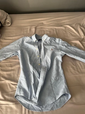 Ljusblå polo skjorta med broderad logga - Klassisk ljusblå skjorta i bomull med button-down krage och broderad logga på bröstet. Skjortan har långa ärmar och vita knappar framtill. Perfekt för en clean och stilren look.