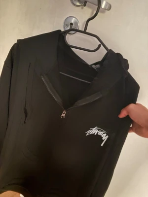 Svart Stüssy hoodie med dragkedja - Svart hoodie från Stüssy med halv dragkedja framtill och vit Stüssy-logga på bröstet. På ryggen finns en stor vit logga och två tärningar. Hoodien har huva och känguruficka. Perfekt för streetwear-stilen. Oversized!