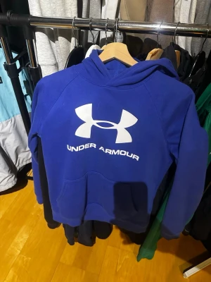 Blå Under Armour hoodie med logga - Snygg blå hoodie från Under Armour med stor vit logga och text på bröstet. Hoodien har huva och magficka, samt är tillverkad i ett mjukt och varmt material, perfekt för kalla dagar. Modellen är loose fit och har ColdGear-teknologi.