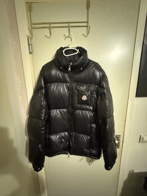 Svart dunjacka från Moncler - Snygg svart dunjacka från Moncler med glansig finish och hög krage. Jackan har en bröstficka med lock och Moncler-logga, dragkedja framtill och stora vadderade paneler. Perfekt för kalla vinterdagar och riktigt trendig look.