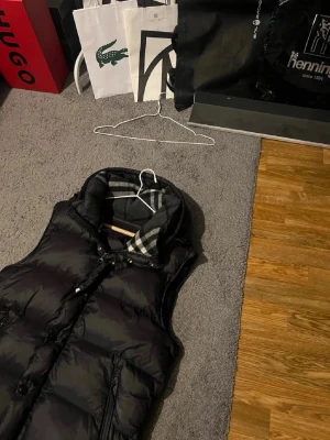 Burberry väst - Snygg svart dunväst med hög krage och dragkedja samt knappar framtill. Insidan av kragen har ett grått och vitt rutigt foder som ger en stilren kontrast. Perfekt för lager-på-lager och håller dig varm under kyliga dagar. Tyvärr saknar den ärmar men fungerar fortfarande bra till vardagliga stunder 