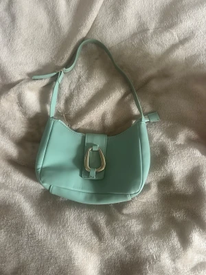 Ljusgrön axelremsväska i skinnimitation - Trendig ljusgrön handväska i skinnimitation med axelrem och silverfärgat spänne framtill. Väskan har en mjuk, rundad form och minimalistisk design som ger en fräsch look. Perfekt accessoar för att lyfta din outfit.