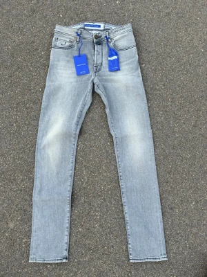 Ljusgrå bard fit jeans Jacob Cohën - Snygga ljusgrå jeans från Jacob Cohën i slim fit-modell. Jeansen har klassisk femficksdesign, subtila slitningar och detaljer som blå etiketter och broderad J på myntfickan. Tillverkade i mjuk denim med stretch för extra komfort.