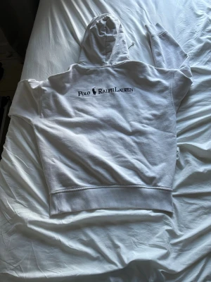 Vit hoodie från Polo Ralph Lauren - Säljer en vit hoodie från Polo Ralph Lauren med klassisk logga på bröstet och stor broderad text på ryggen. Tillverkad i mjukt bomullsmaterial och har ribbade muddar vid ärmslut och nederkant. Kostade 3000 kr ny, mitt pris 1700