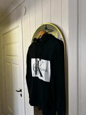 Svart hoodie från Calvin Klein Jeans - Svart hoodie från Calvin Klein Jeans med stor vit logga på bröstet. Klassisk huva med dragsko och mjukt material som känns skönt mot huden. Perfekt för dig som gillar stilrena och tidlösa plagg med en modern touch.