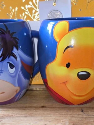 Disney Tams Nalle Puh och Ior muggar - Två färgglada muggar med motiv av Nalle Puh och Ior från Disney. Muggarna är blå med stora tryck av karaktärernas ansikten och har rundad form med bekvämt handtag. Perfekta för te eller varm choklad och ger en nostalgisk känsla till fikastunden. Höjd 11,5 cm, Diam 8,5 cm